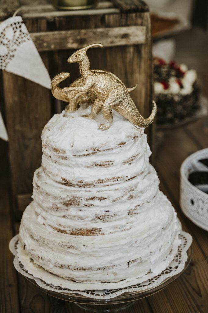 Mehrstöckiger Kuchen mit weißen Zuckerguss und goldenen Dinosaurierfiguren obenauf.
