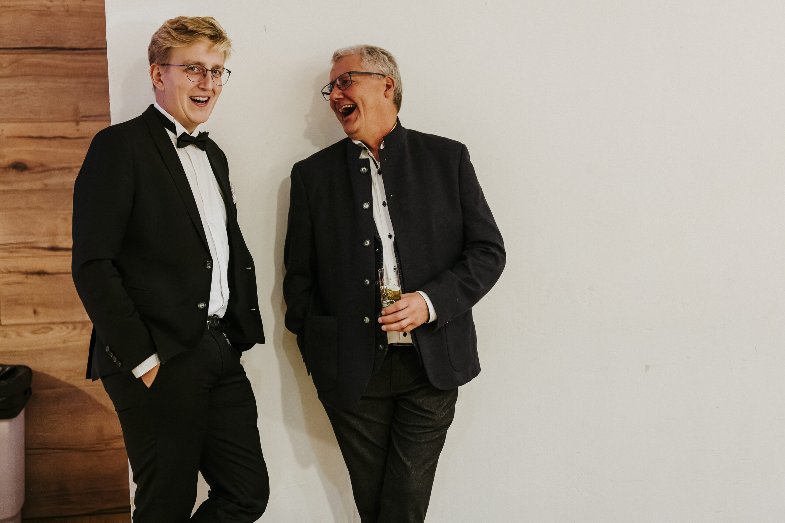 Two men laughing, one in a tuxedo and the other in a dark jacket holding a drink.