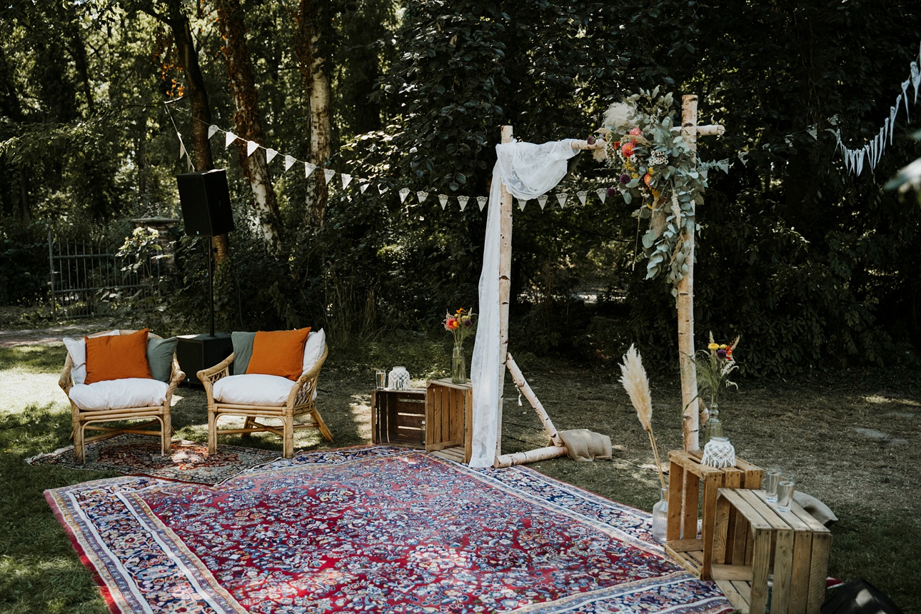 Outdoor-Sitzbereich im Wald mit Stühlen, Bögen, Blumen, Holzkisten und Wimpelketten.