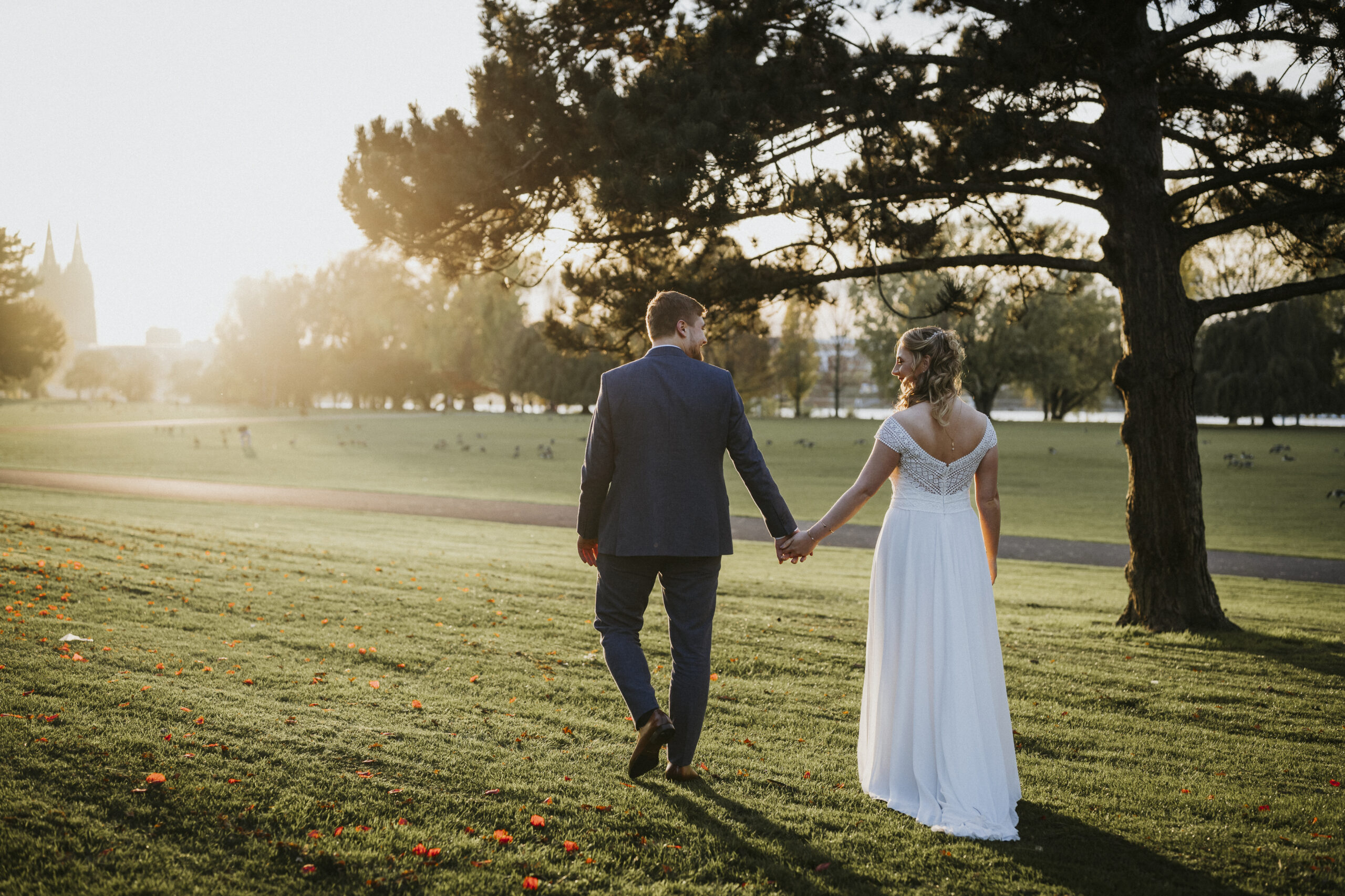 Paar in eleganter Kleidung spaziert bei Sonnenuntergang Hand in Hand über Wiese mit Bäumen.
