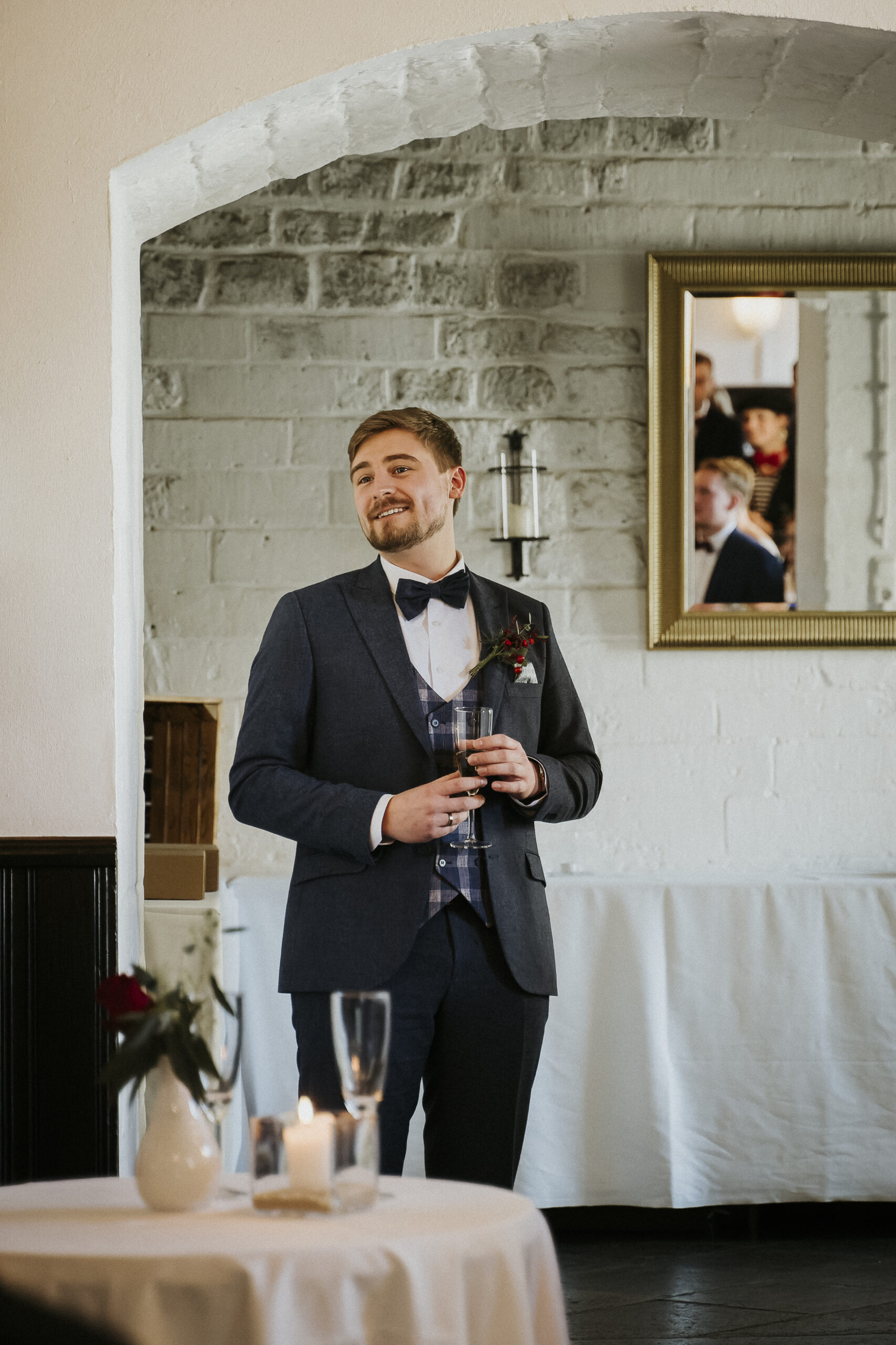 Person im Anzug mit Glas, stehender festlicher Raum, Spiegel, Tisch mit Kerze und Blumen.