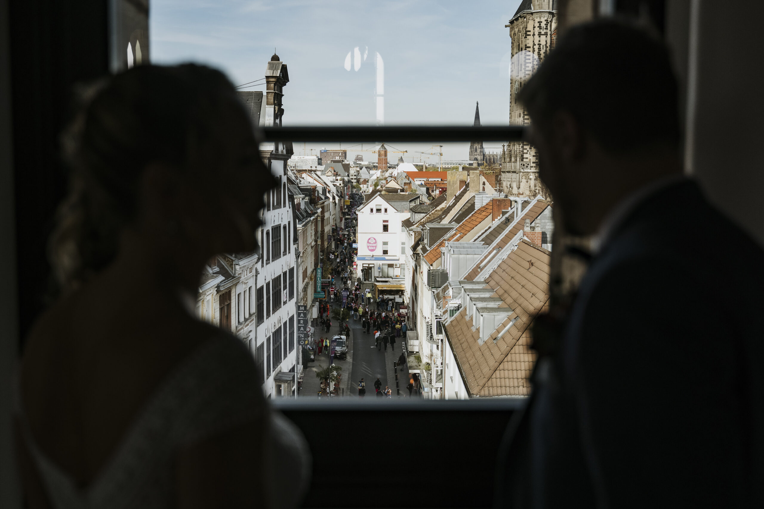 Zwei Silhouetten blicken von einem Fenster auf eine belebte Straße mit historischen Gebäuden.