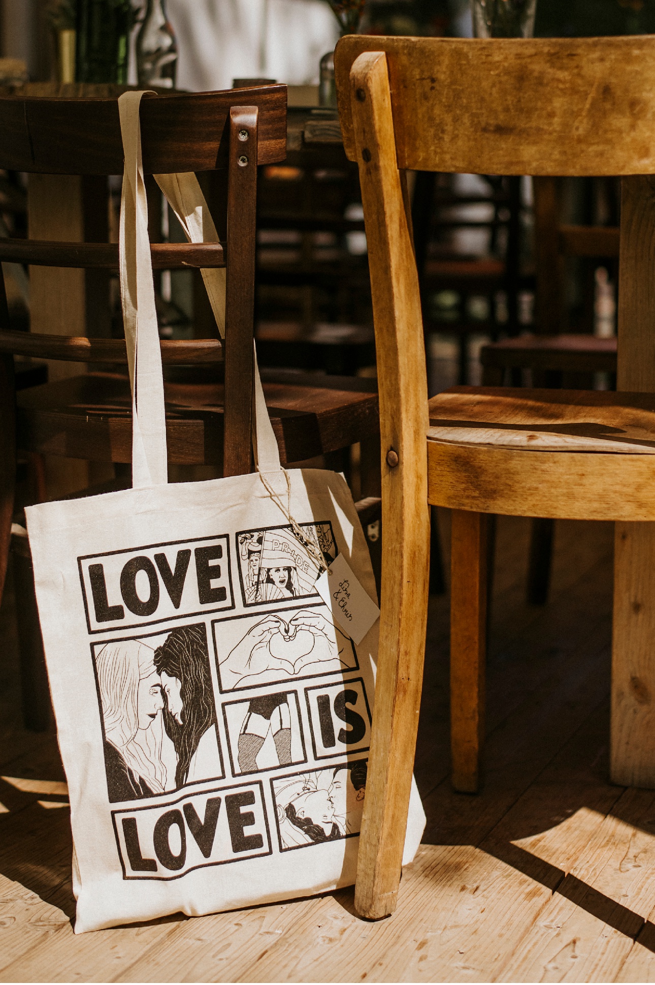 Stofftasche mit "LOVE IS LOVE"-Aufdruck und Illustrationen an einem Holzstuhl.