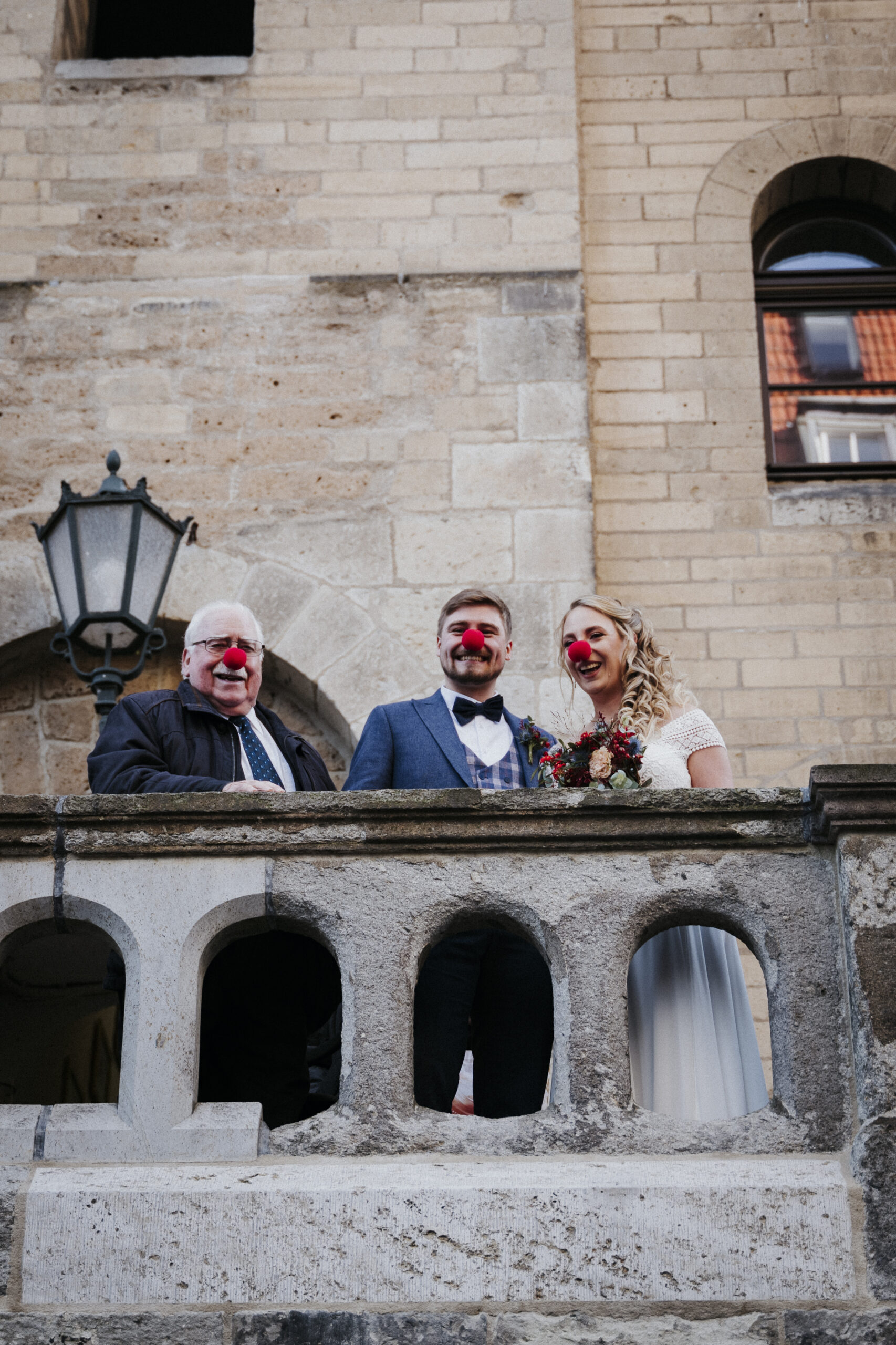 Drei Personen mit roten Clownsnasen auf einem Balkon vor einem historischen Gebäude.