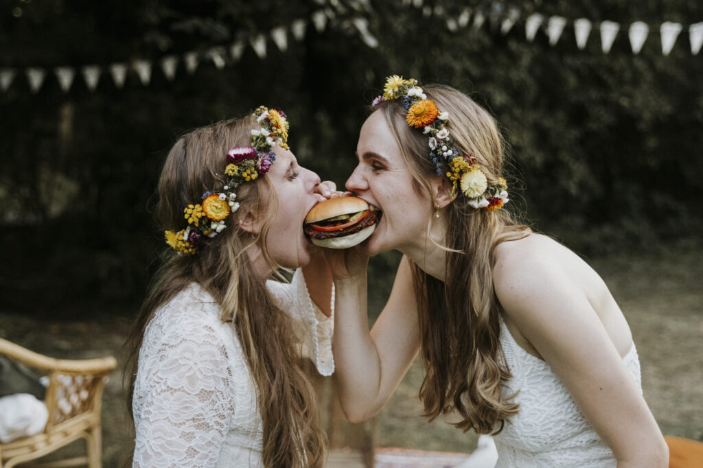 Two people in white dresses with flower crowns share a burger outdoors.