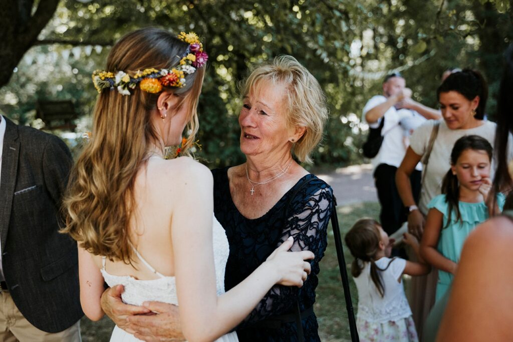 Frau im weißen Kleid mit Blumenkranz wird von älterer Frau im dunklen Kleid umarmt.