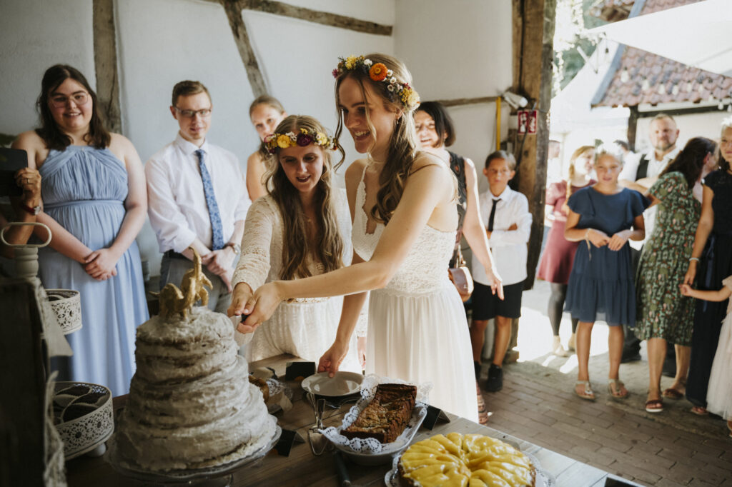 Two women in white dresses cut a wedding cake surrounded by people in festive attire.