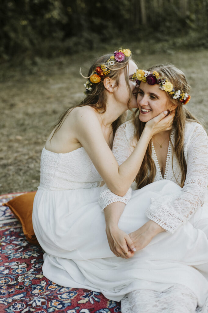 Zwei Frauen in weißen Spitzenkleidern und Blumenkränzen flüstern und lächeln im Freien.