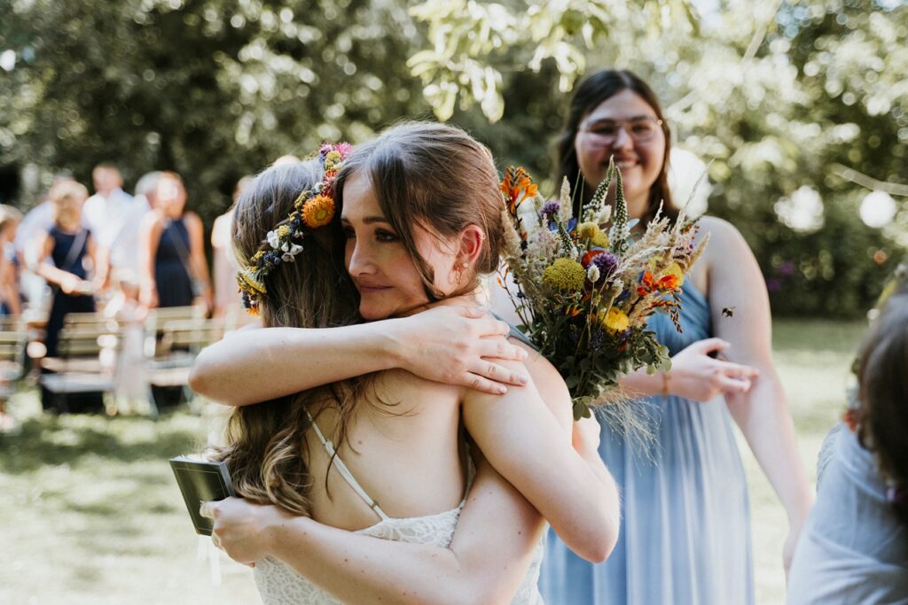 Zwei Frauen umarmen sich im Freien, eine hält einen Blumenstrauß, umgeben von Gästen im Park.