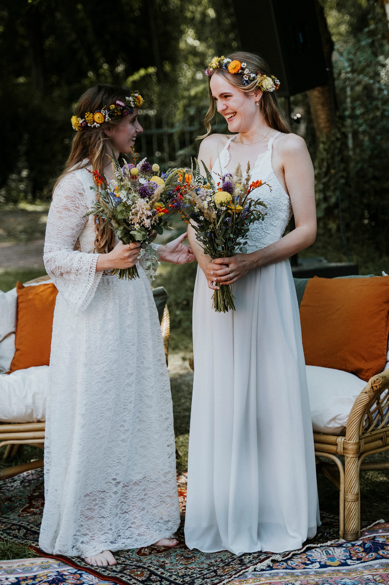 Zwei Frauen in weißen Kleidern mit Blumenkränzen halten Blumensträuße auf buntem Teppich im Freien.