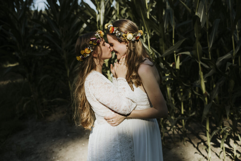 Zwei Personen in weißen Kleidern mit Blumenkränzen im Haar in einem Feld hoher Pflanzen.