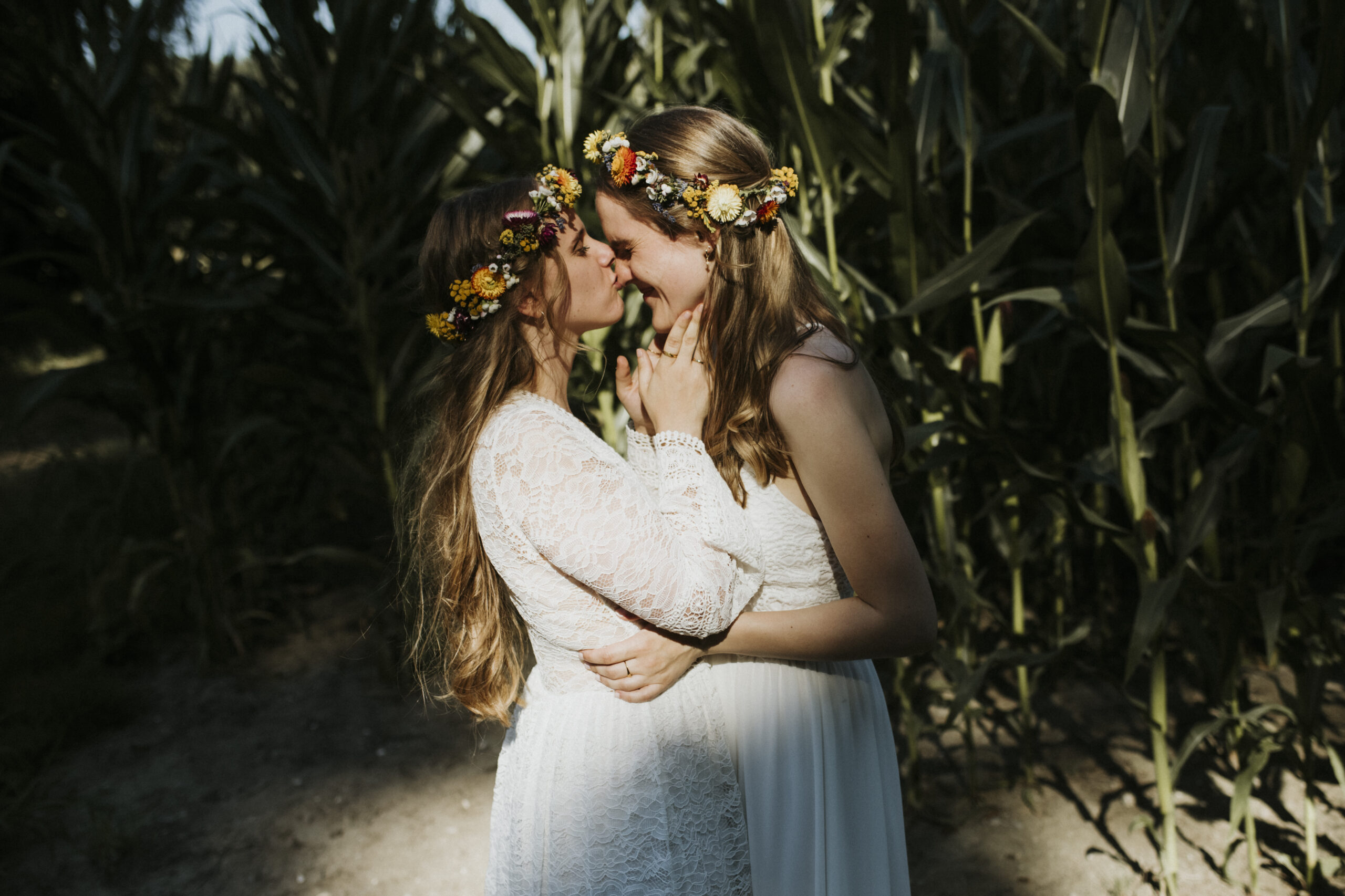 Zwei Personen in weißen Kleidern mit Blumenkränzen im Haar in einem Feld hoher Pflanzen.