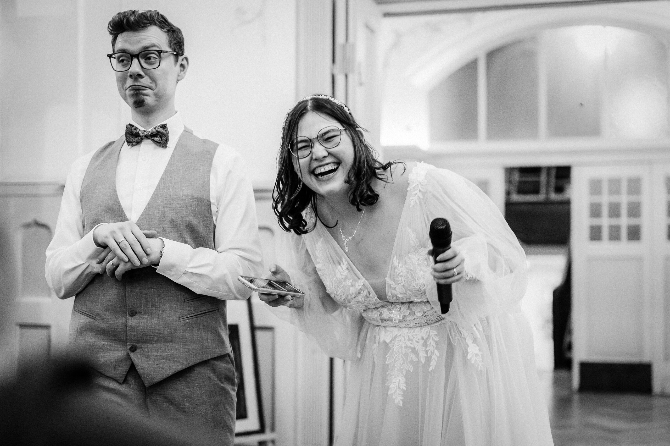 Braut mit Mikrofon lacht während einer Hochzeitsrede, daneben steht der Bräutigam und reagiert humorvoll – dokumentarische Hochzeitsfotografie in Schwarzweiß.
