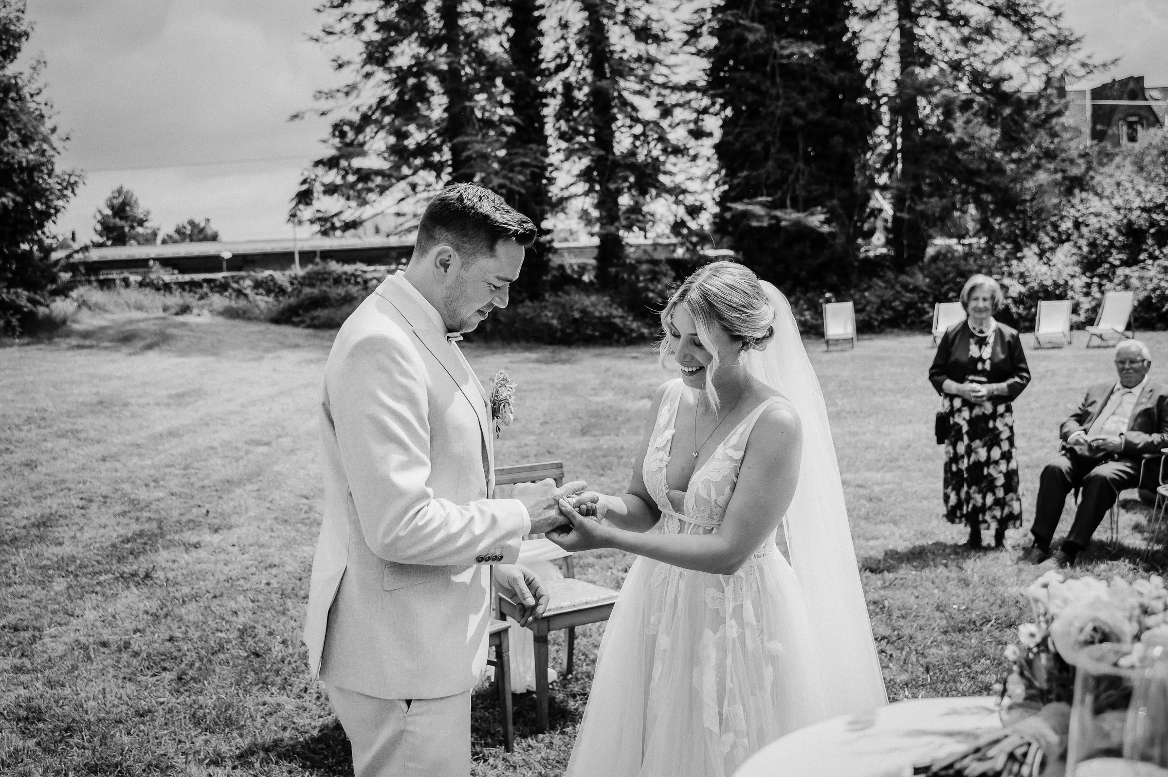 Braut und Bräutigam bei der Zeremonie im Garten, Handschuhwechsel während der Hochzeit, im Hintergrund Gäste sitzend.