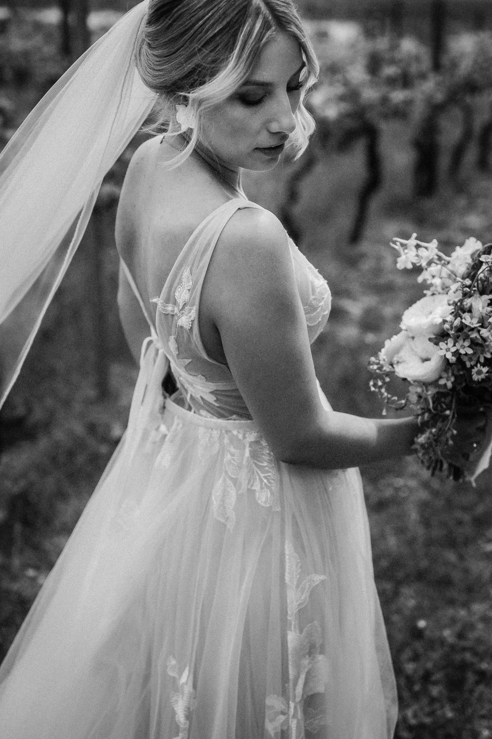 Braut in Hochzeitskleid mit Schleier, seitlich fotografiert, hält Blumenstrauß außerhalb des Bildes.