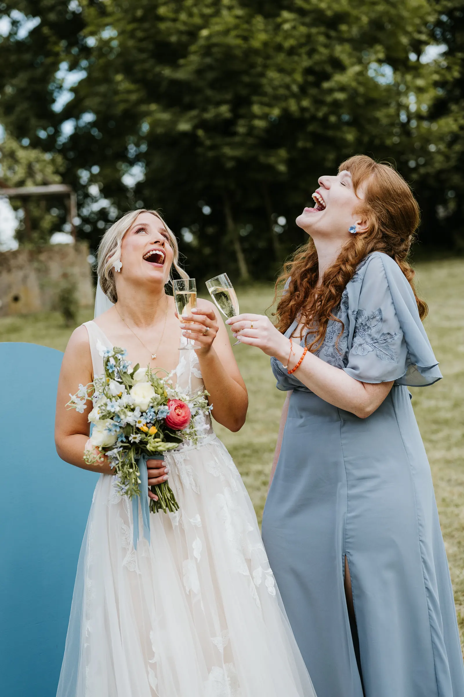 Braut in weißem Kleid und Brautjungfer lachen, stoßen mit Sekt an, draußen mit Blumenstrauß.