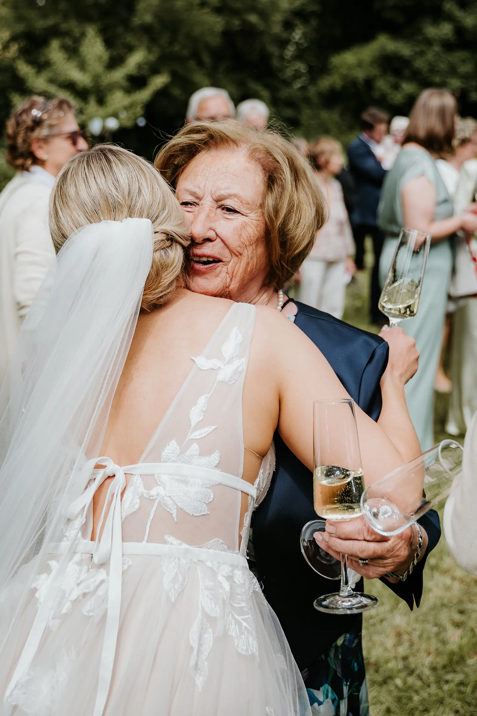 Braut in weißem Brautkleid umarmt eine ältere Frau, feiert mit Sektgläsern im Freien.