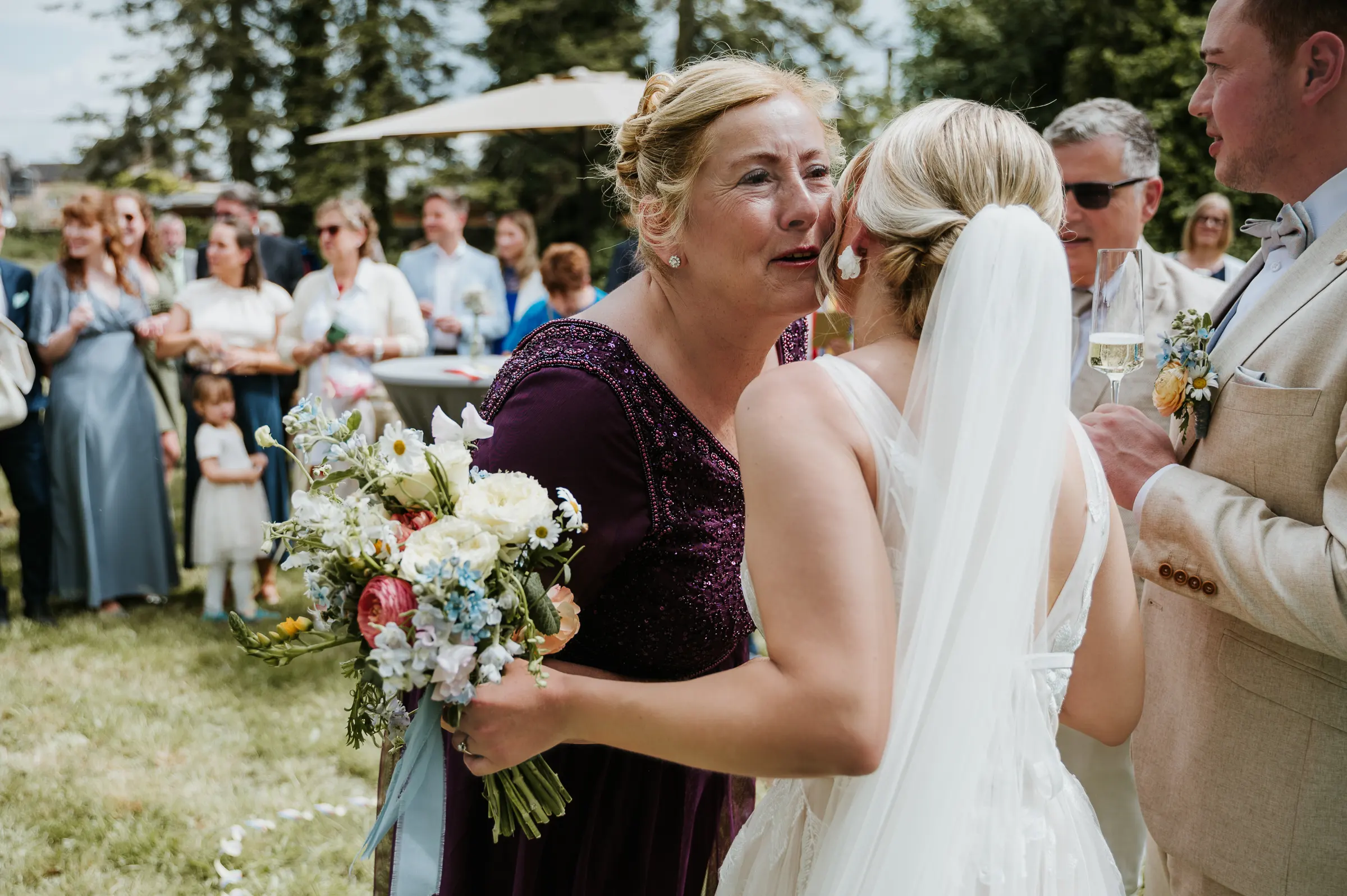 Braut in Weiß umarmt von Frau in dunklem Glitzerkleid, Braut mit Blumenstrauß, Hochzeitsfeier im Freien.