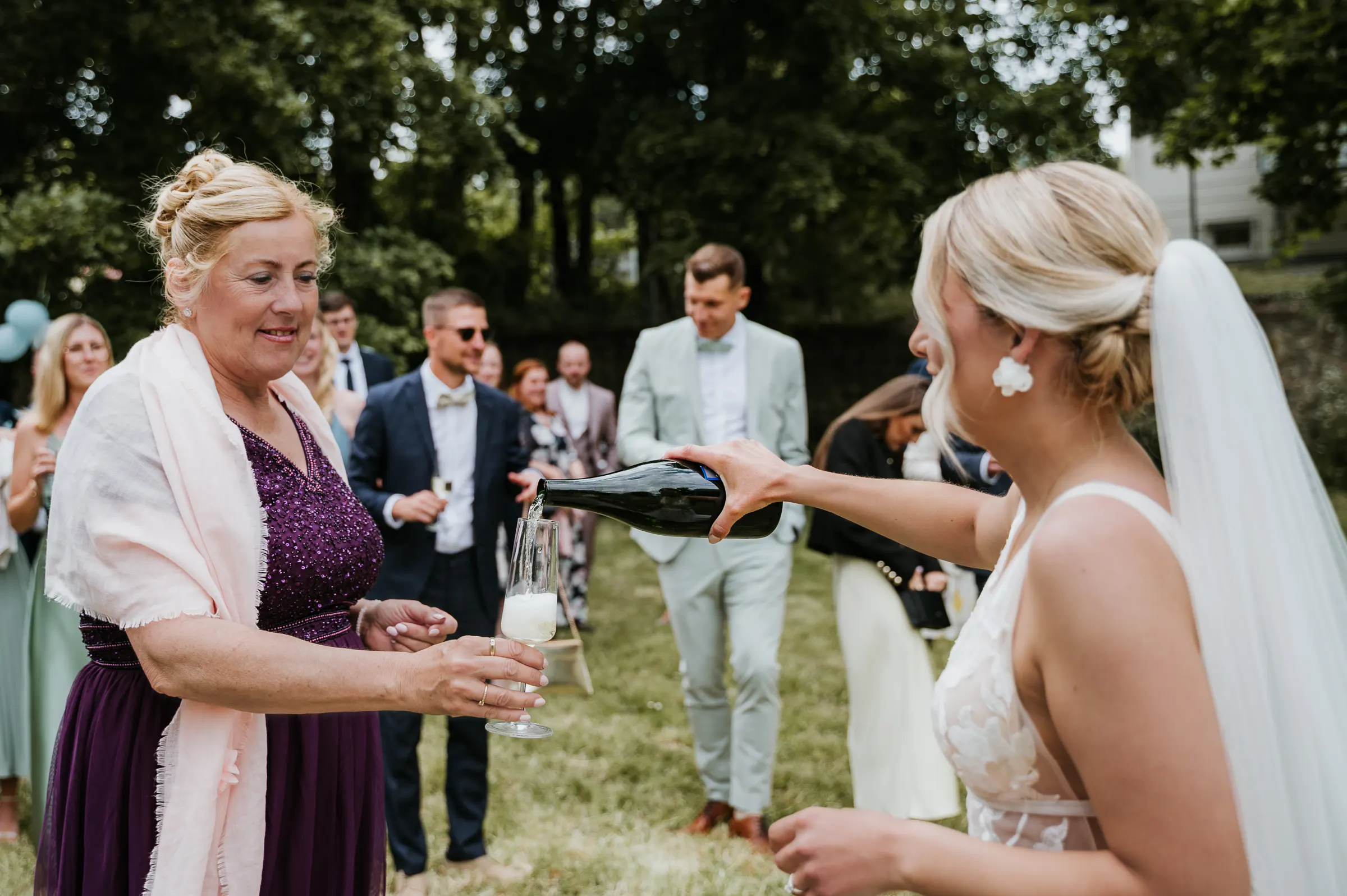 Braut schenkt Glas Schaumwein aus Flasche zu Frau in violettem Kleid bei Outdoor-Hochzeit, Gäste im Hintergrund.