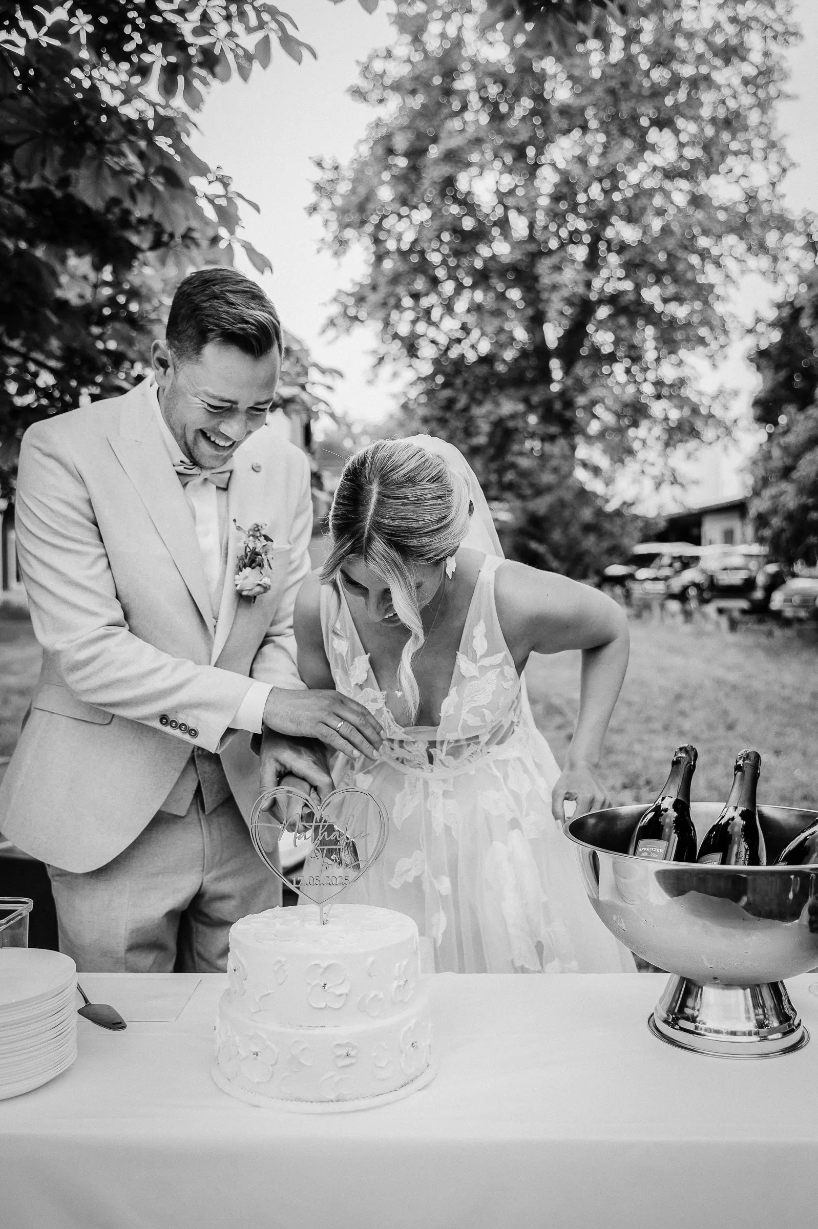 Braut und Bräutigam schneiden Kuchen bei Hochzeit im Freien; Heißgetränke im Eiskübel neben einem Herz-Topper.