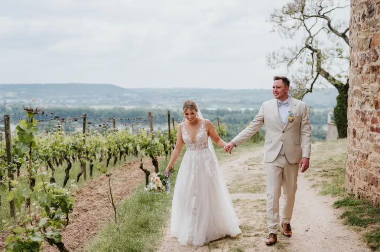 Braut und Bräutigam gehen Hand in Hand durch Weinberge mit Blick über Landschaft; Braut in weißem Kleid, Bräutigam im hellen Anzug.