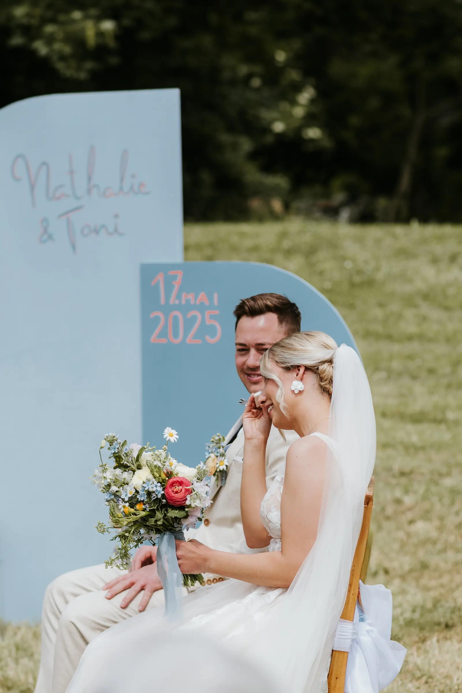 Brautpaar mit Blumen sitzt draußen neben hellblauem Hochzeitsbrett und Datumslog.