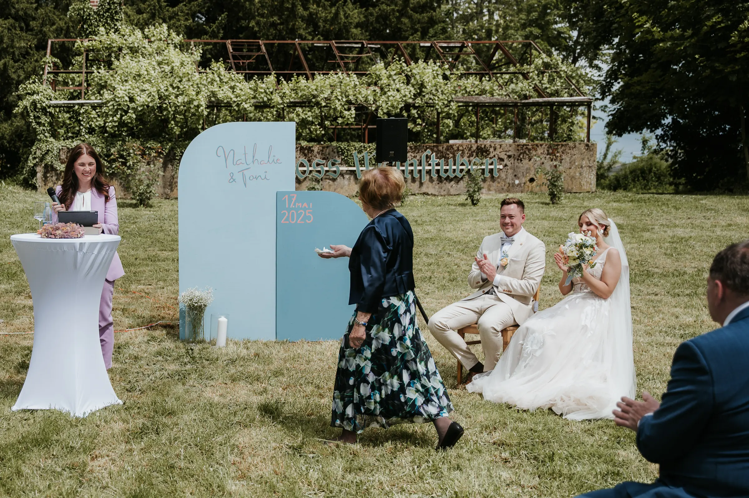 Brautpaar sitzt während einer Outdoor-Hochzeit, Zeremoniebereich mit blauen Schildern und Blumen.