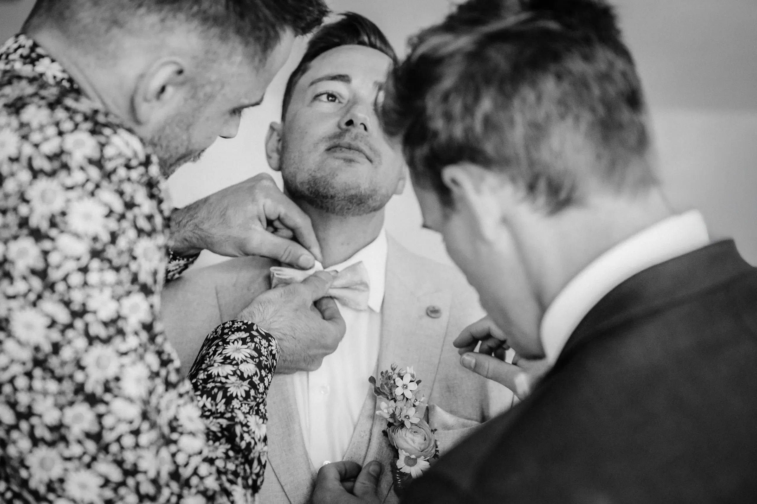 Drei Männer helfen Mann im Anzug beim Aufsetzen der Fliege; einer befestigt eine Blume am Revers. Schwarz-Weiß.