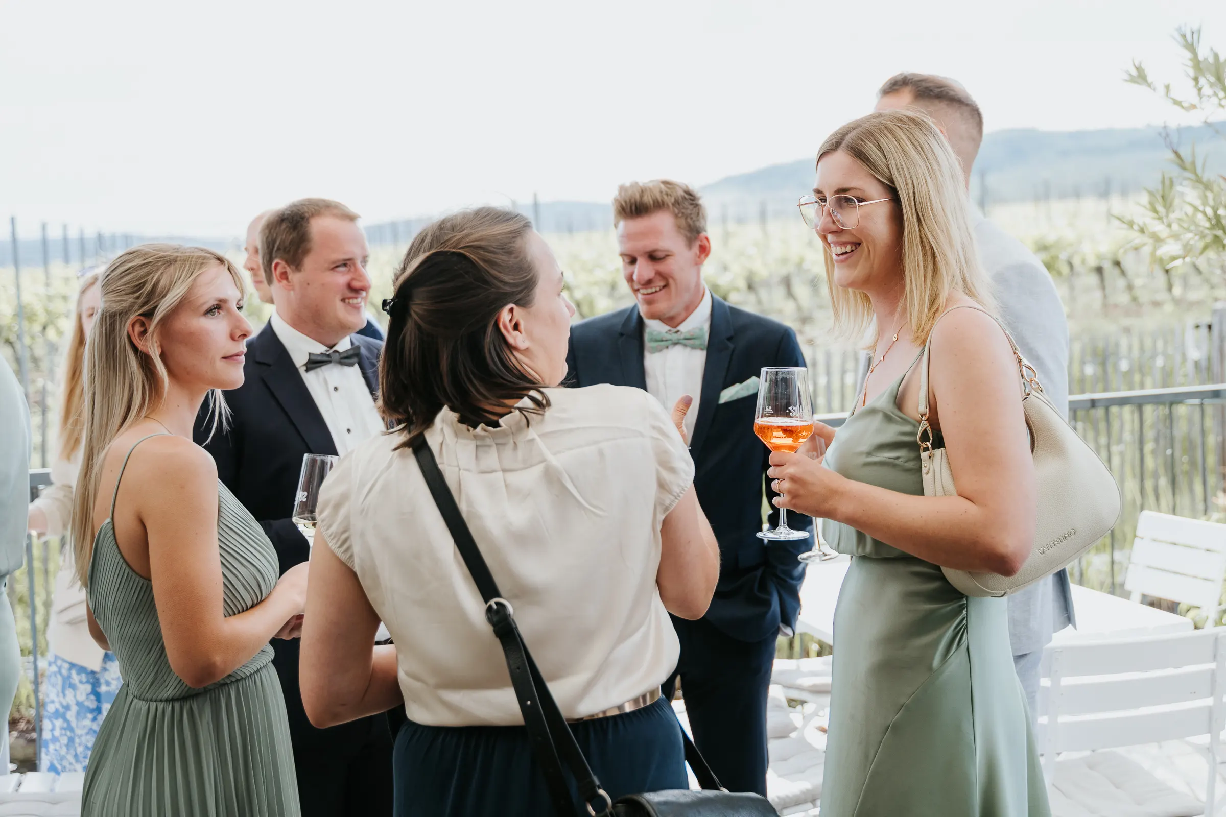 Gruppe elegant gekleideter Menschen bei Outdoors-Event, Gespräche, Gläser mit Wein, Weinberg im Hintergrund