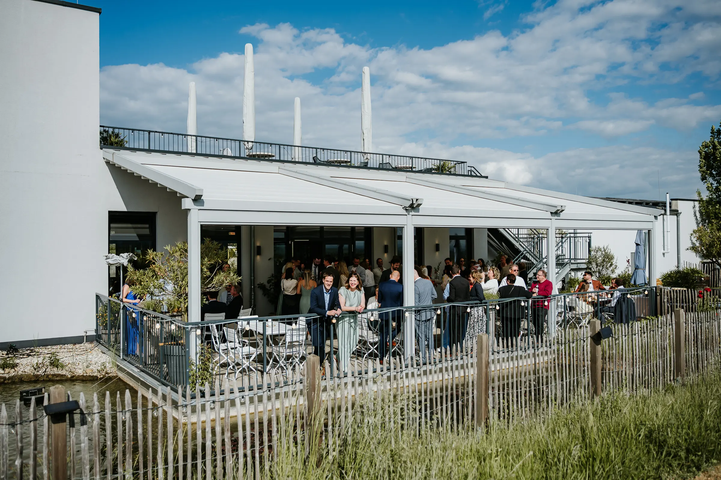 Menschen feiern auf Terrasse eines weißen Gebäudes mit Gartenpflanzen und städtischem Himmel.