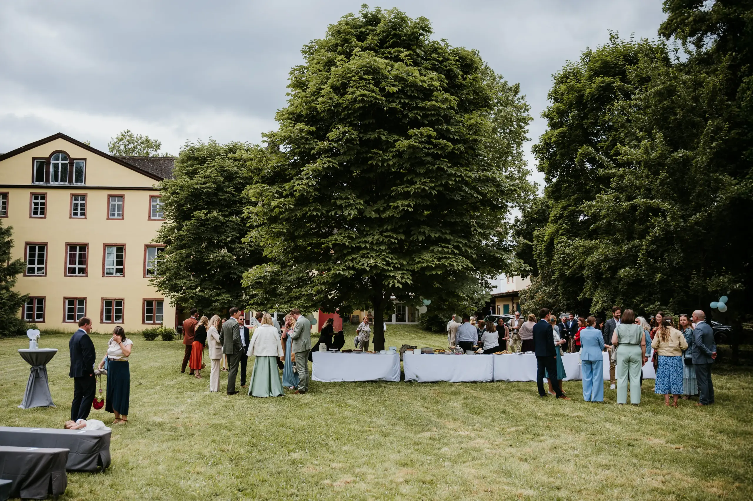 Gruppen von Menschen im Freien bei einer Feier auf grünem Rasen, Buffets und Stehtische, umgeben von Bäumen und einem gelben Gebäudekomplex.