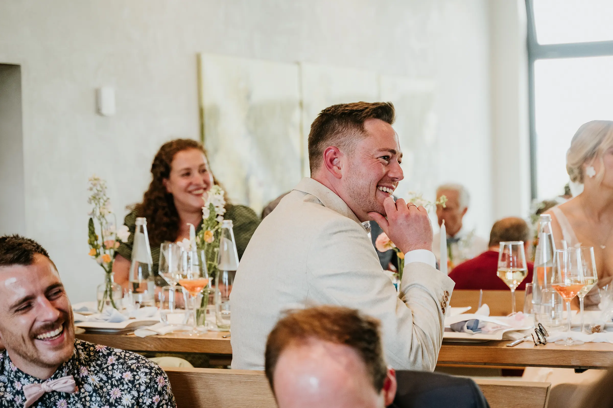 Menschen lachen bei festlicher Tafel mit Blumen, Wein und Gläsern.