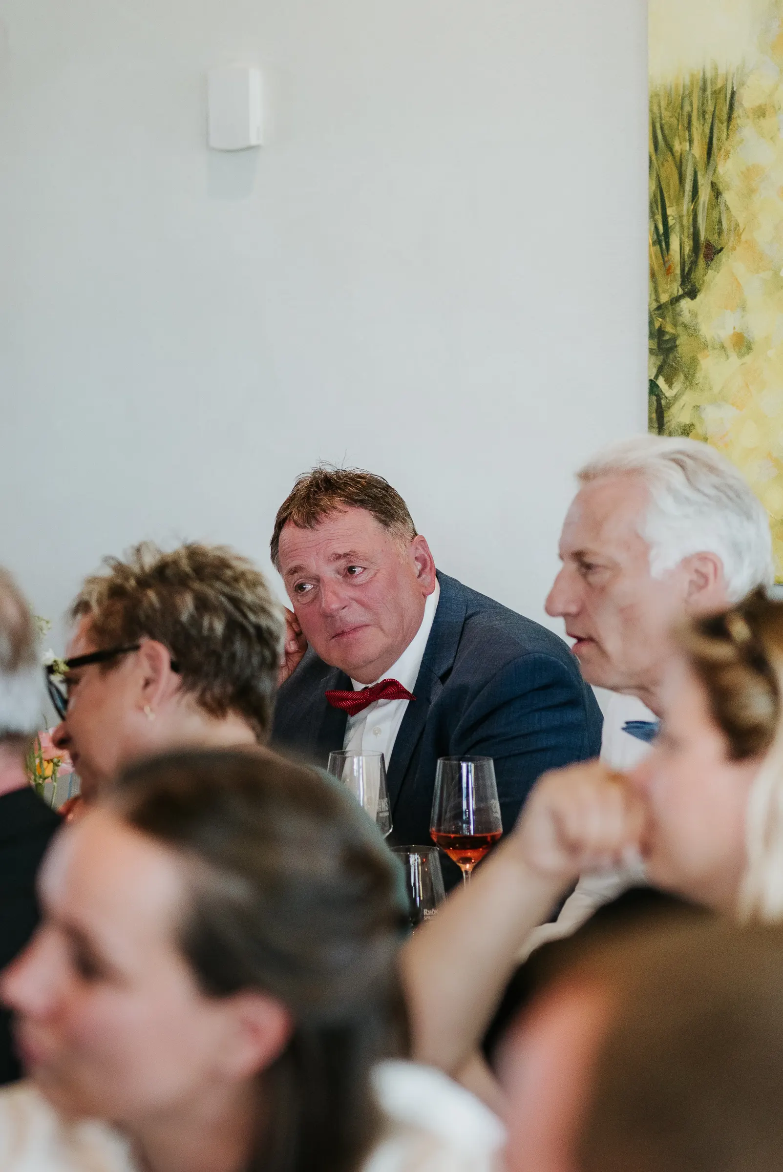 Gruppe von Menschen bei formellem Anlass, Mann mit blauem Anzug und roter Fliege am Tisch, Gläser Wein im Vordergrund.