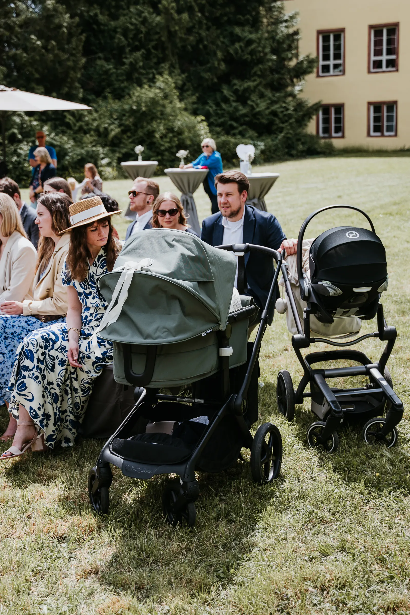Gruppe von Menschen im Freien bei Veranstaltung; vorne Kinderwagen, Stuhlreihen, Gebäude im Hintergrund.