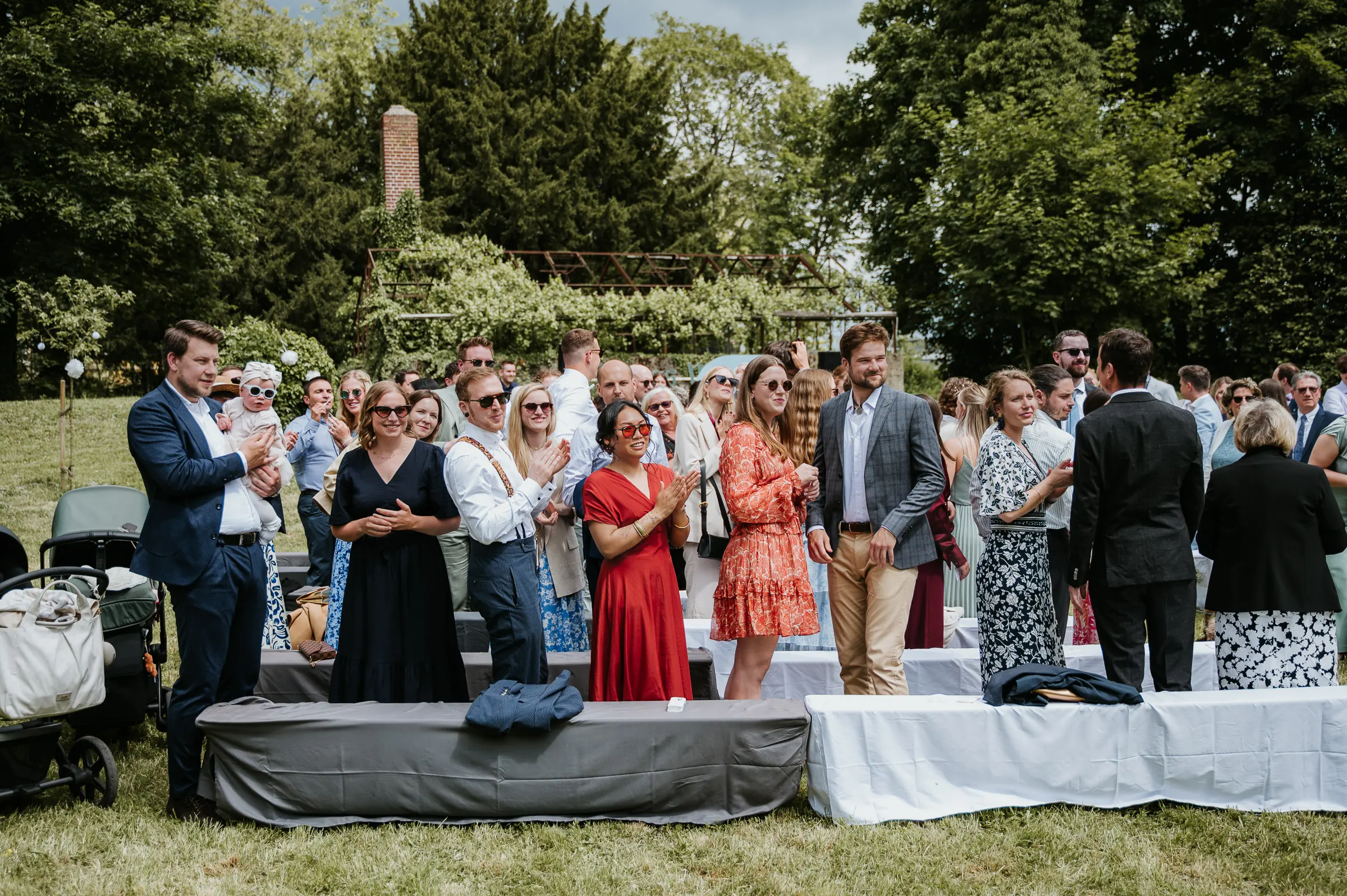 Gruppe gut gekleideter Gäste bei outdoor Zeremonie, Frau in rotem Kleid, Menschen applaudieren, Garten mit Bäumen.