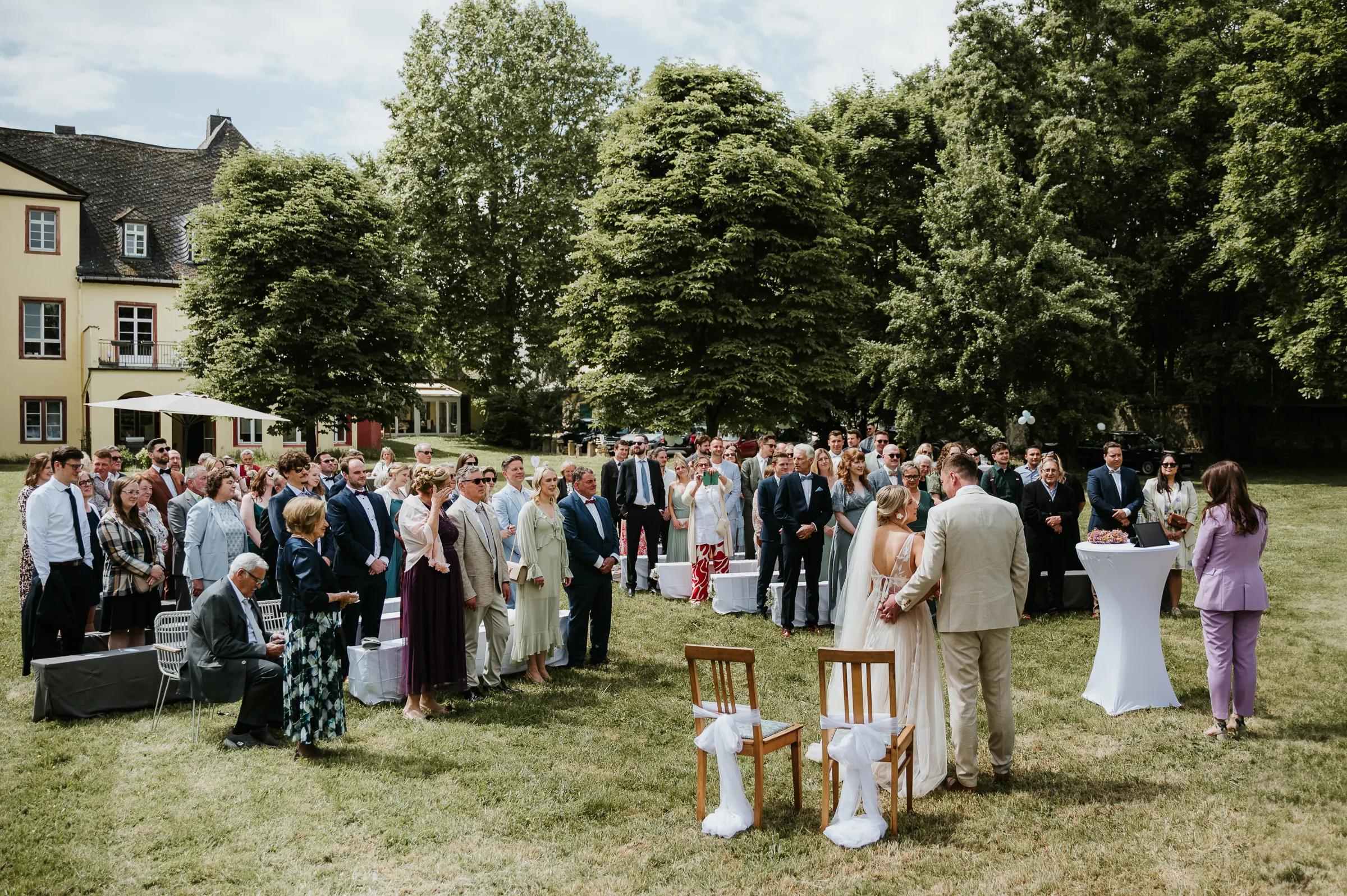Hochzeit im Freien: Gruppe von Gästen auf dem Rasen vor gelb-beigem Gebäude, mit Bäumen im Hintergrund.