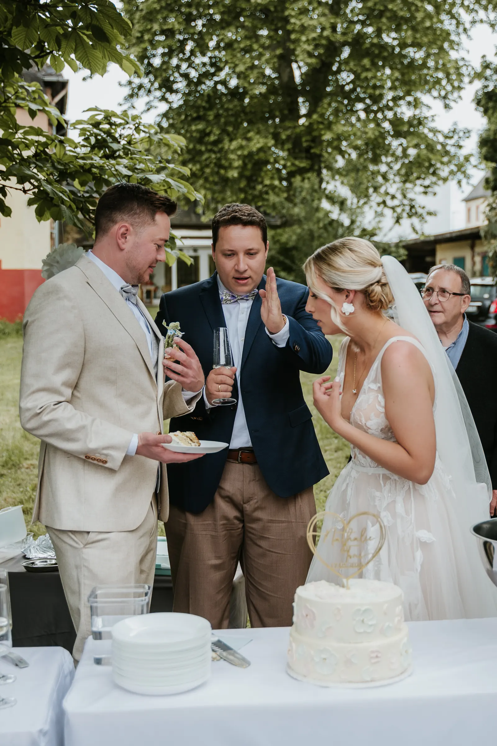 Hochzeitsfeier im Freien: Braut und Bräutigam mit Gästen um einen Kuchen und Torte, Teller und Gläser auf dem Tisch.