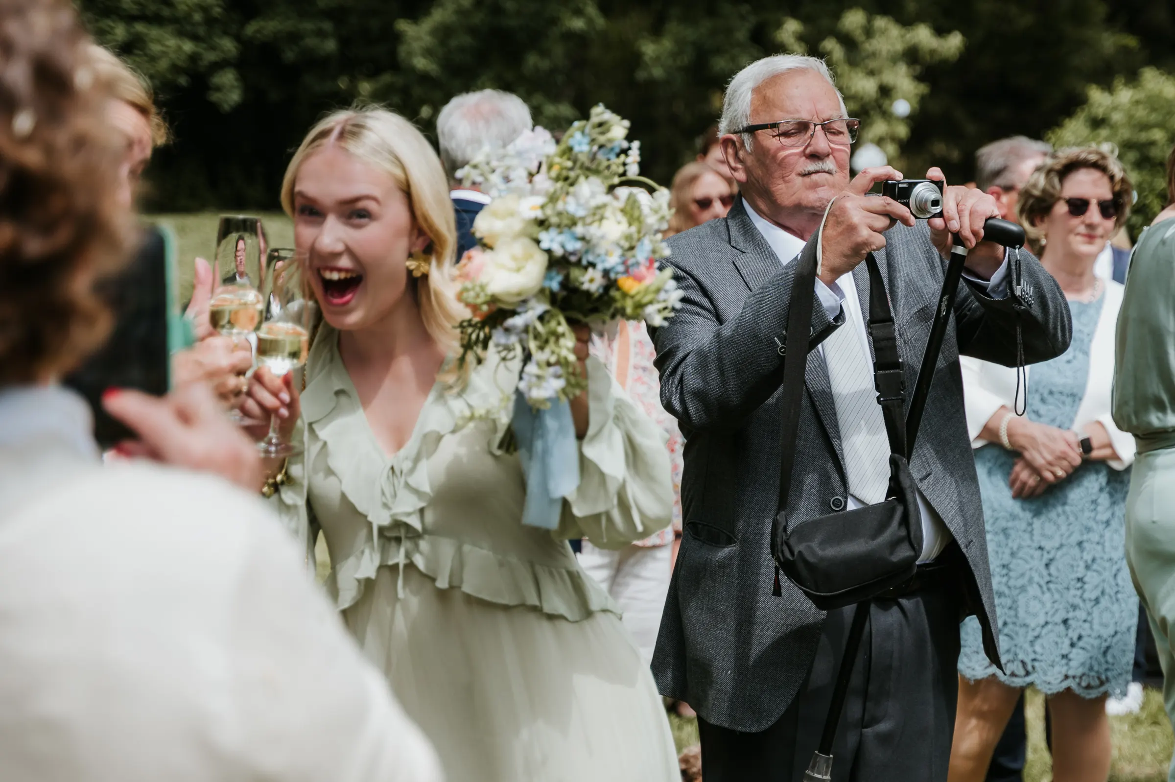 Junge Frau mit Blumenstrauß lacht, hält Gläser, während ein älterer Mann Fotos mit Kamera macht; feiernde Menge im Freien.
