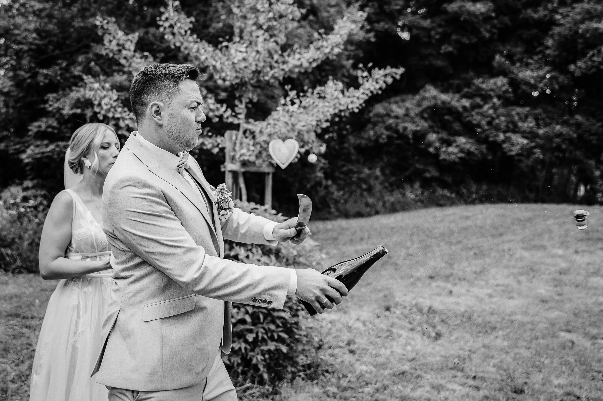 Mann in hellgrauem Anzug hält ein Messer, Braut steht dahinter im Garten, schwarz-weiß Foto.