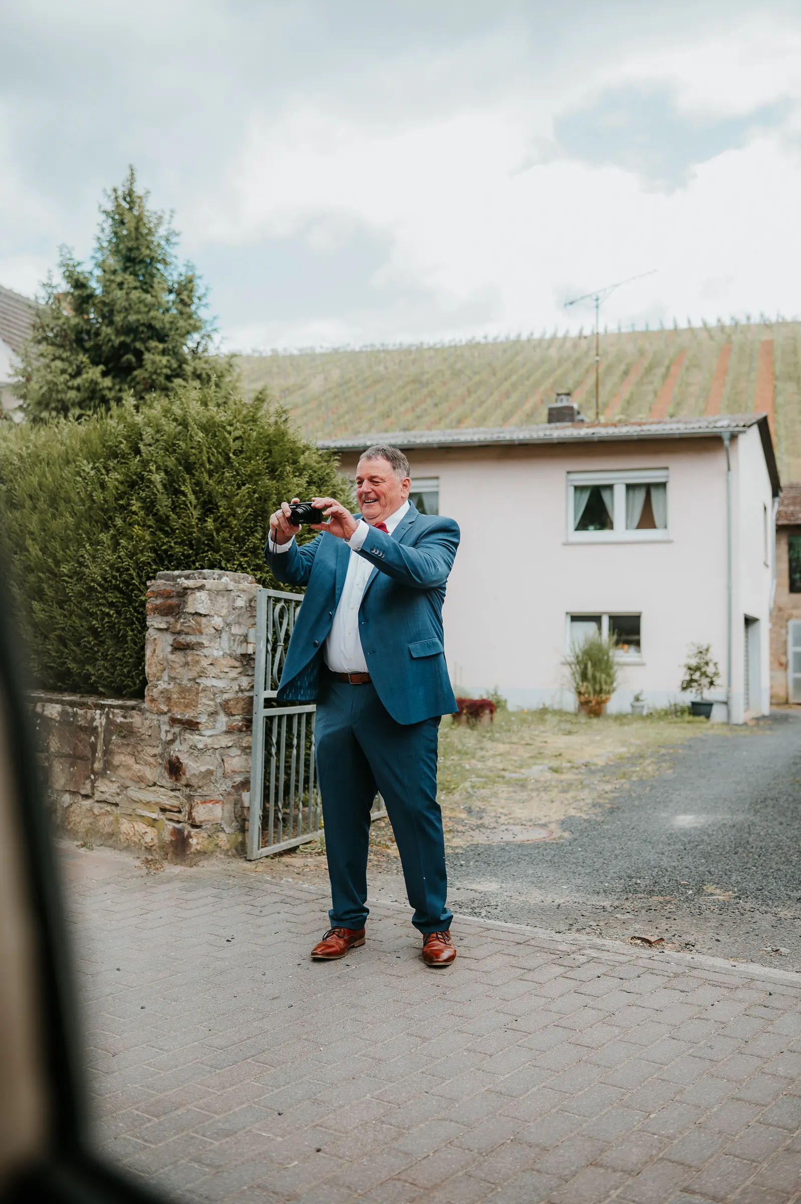 Ein Mann in blauem Anzug fotografiert draußen, vor einem Haus mit Garten, Mauer und Tür.