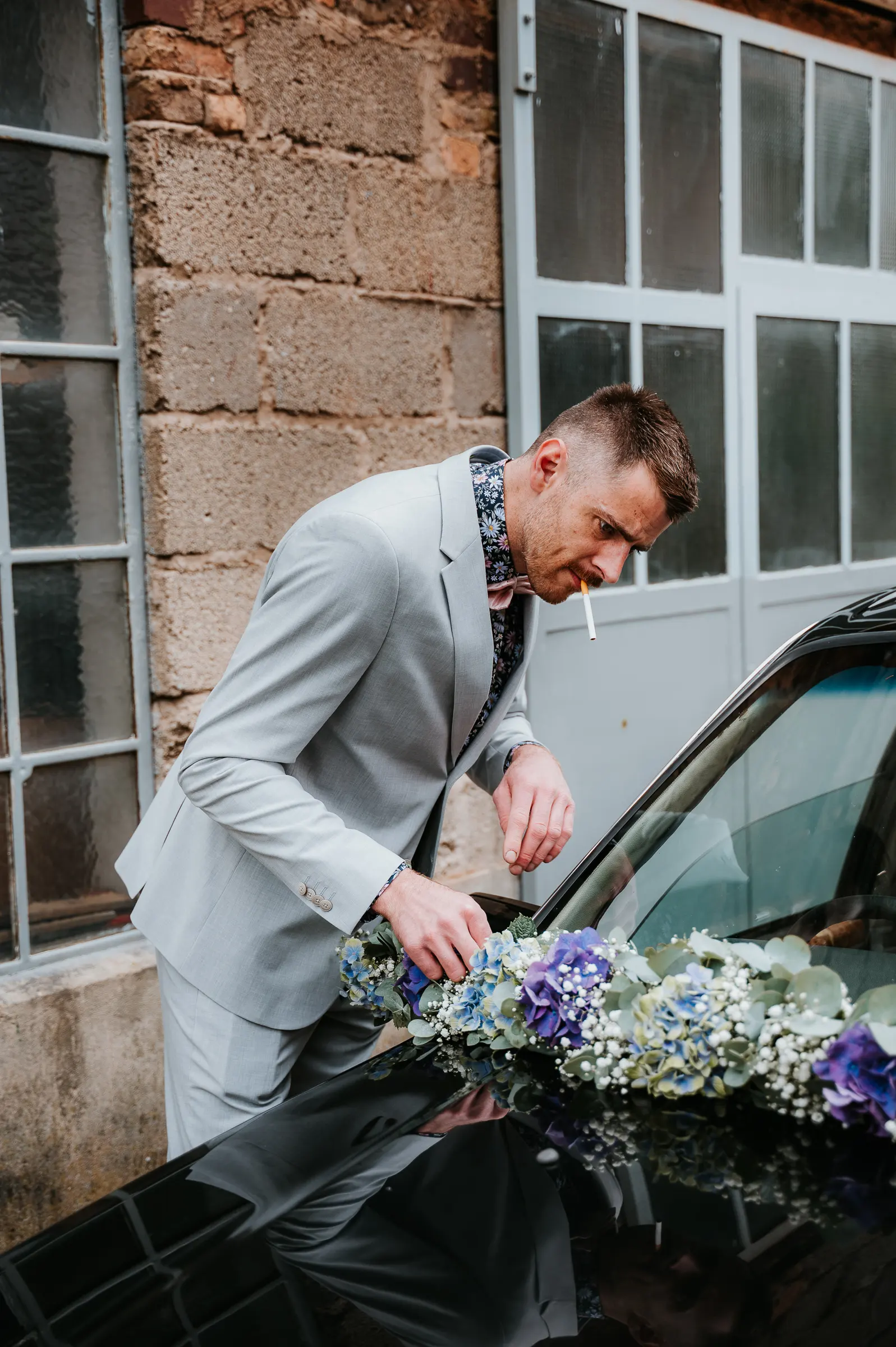 Mann in hellgrauem Anzug legt Blumenkranz auf Autoscheinwerfer, raucht Zigarette.