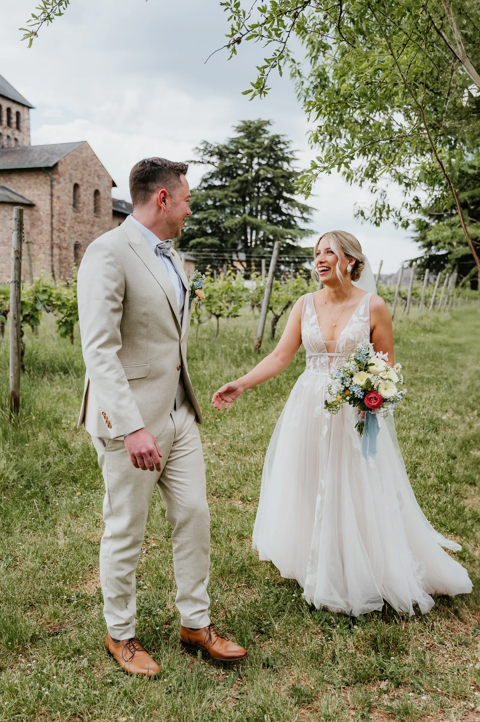 Paar im Hochzeitsoutfit lacht sich in Weinbergumgebung an, holden Händen.