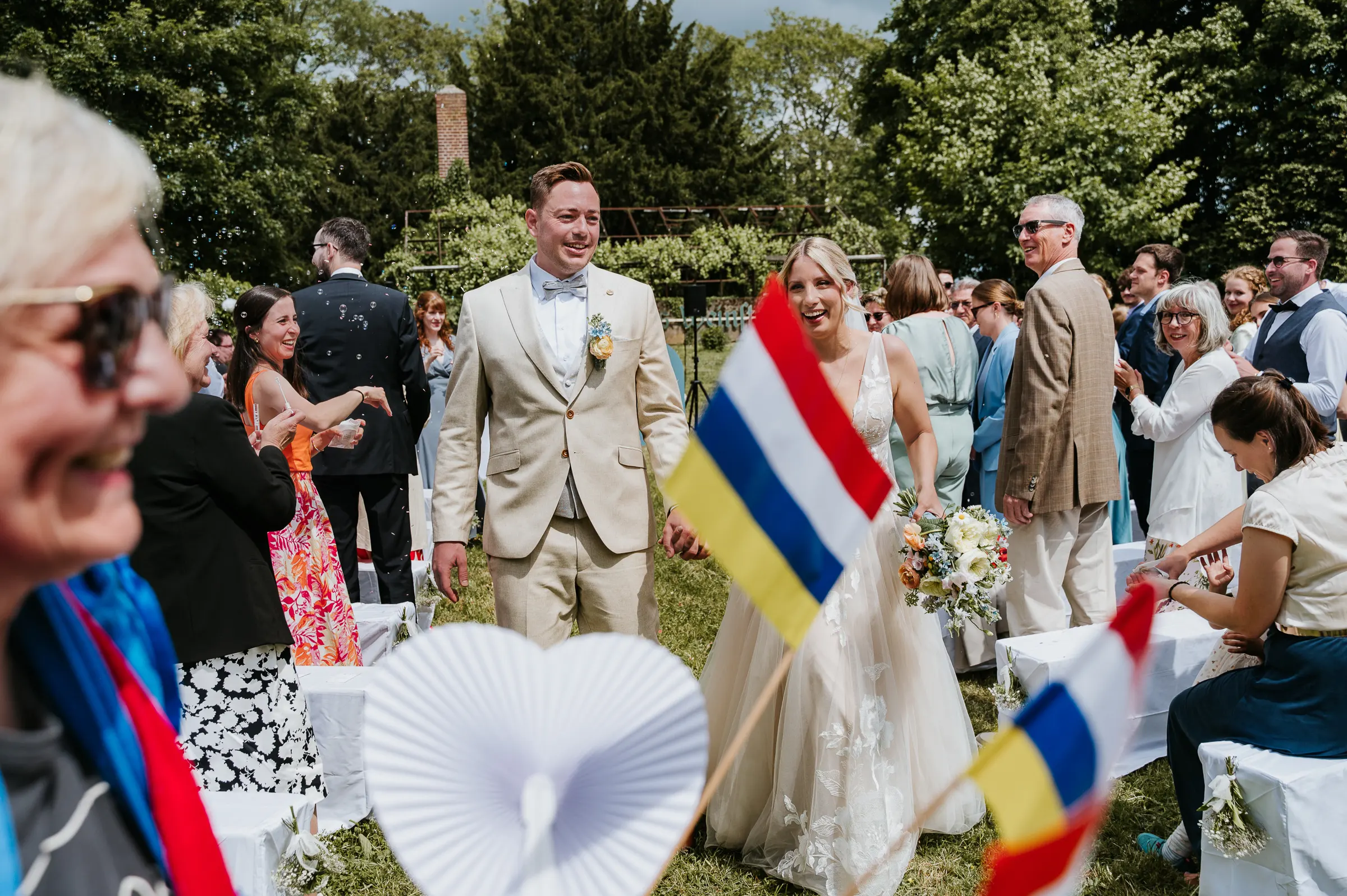 Brautpaar geht lächelnd durch Gäste bei sonniger Outdoor-Hochzeit; Flaggen im Vordergrund, Seifenblasen, Bäume und Himmel im Hintergrund.