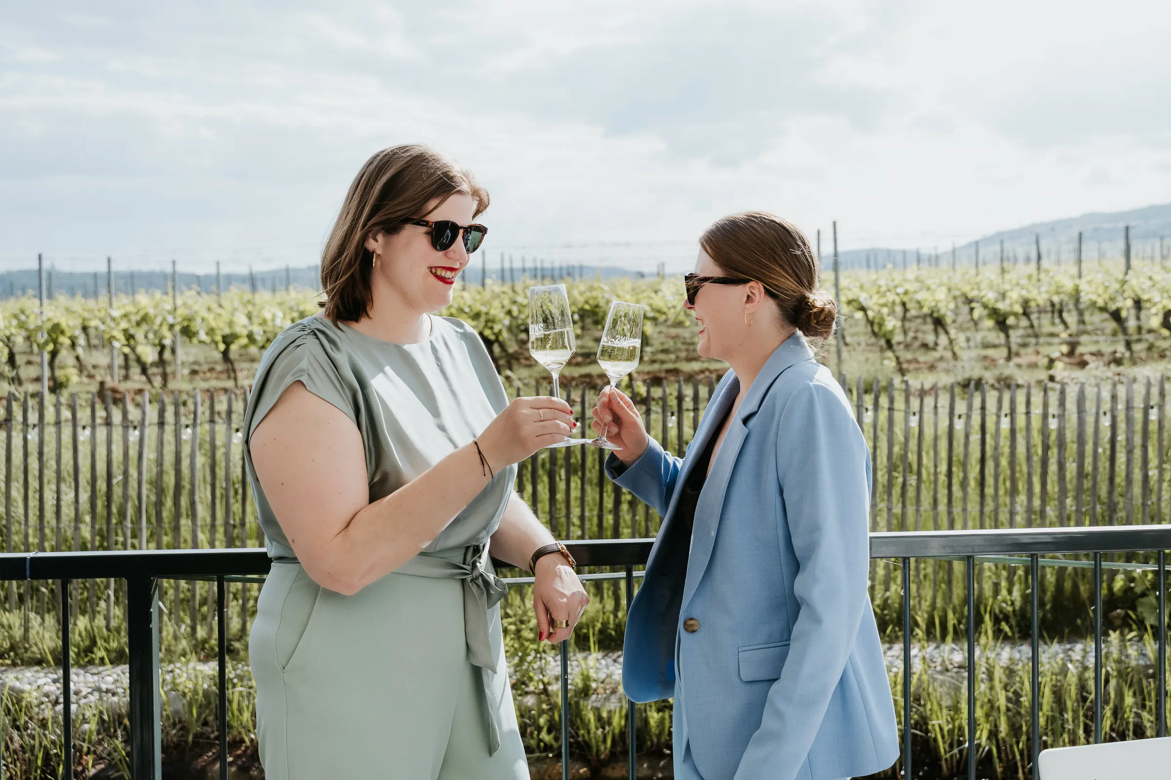 Two women đứng ngoài, stoßen mit Sektgläsern an, lächeln vor Weinbergen, tragen Sonnenbrillen.