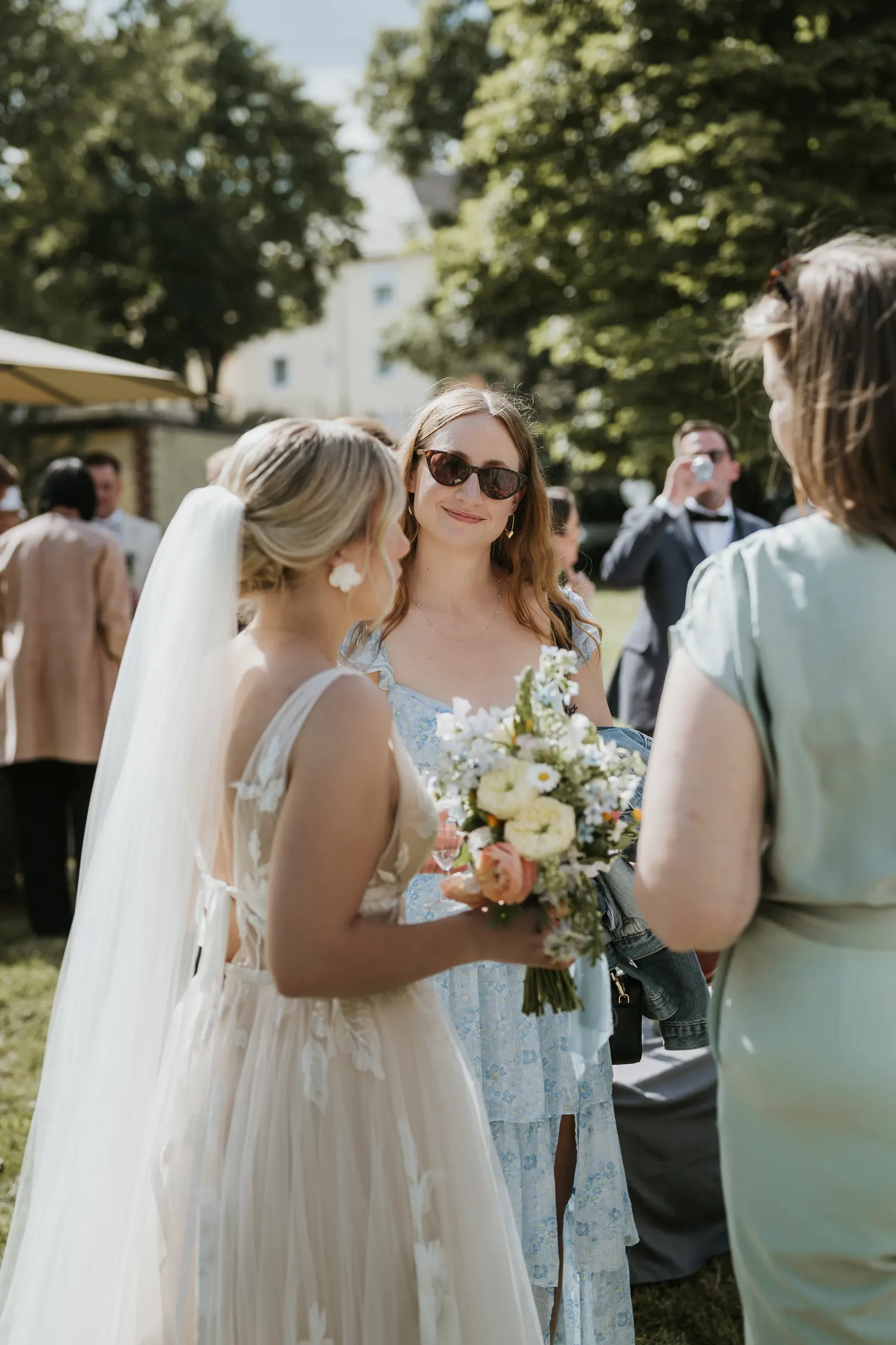 Zwei Hochzeitsgäste im Freien, eine Braut mit Schleier und Blumen, eine weitere Frau mit Blumenstrauß, Bildmitte mit Sonnenbrille.